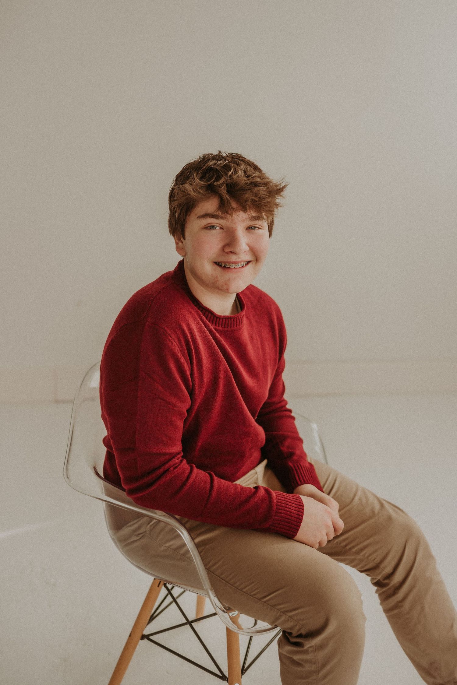 Young person in red sweater smiles while sitting on modern chair in studio setting.