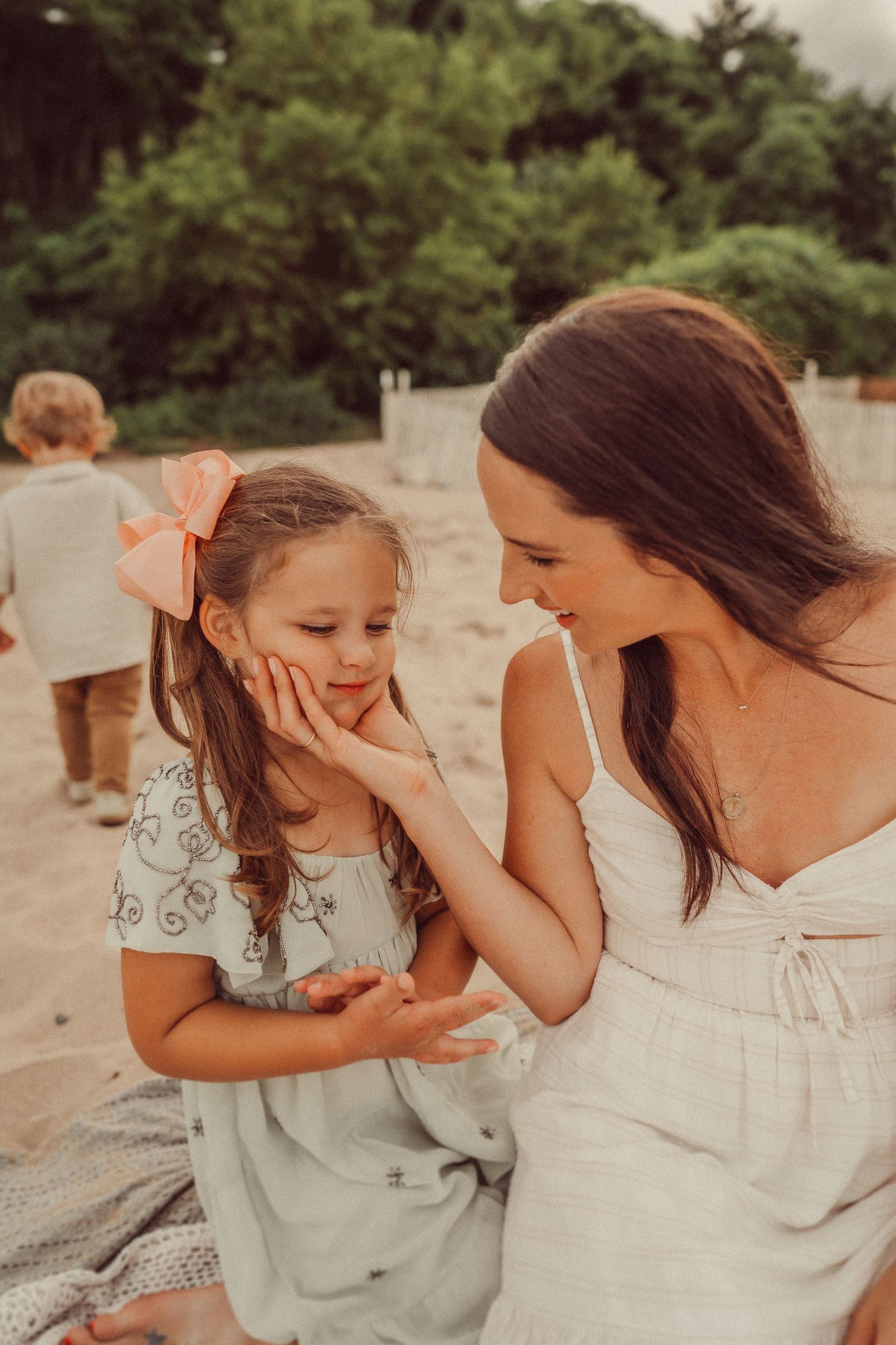 Two people in white dresses share a tender moment while sitting on sand at the beach.