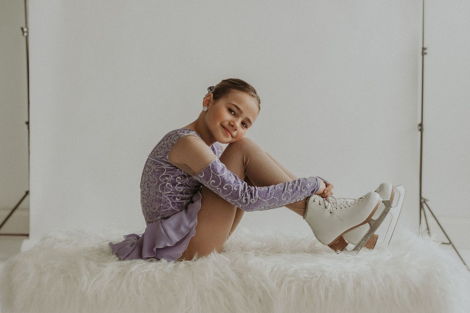 Figure skater in purple dress relaxes on white fur surface while adjusting ice skates.