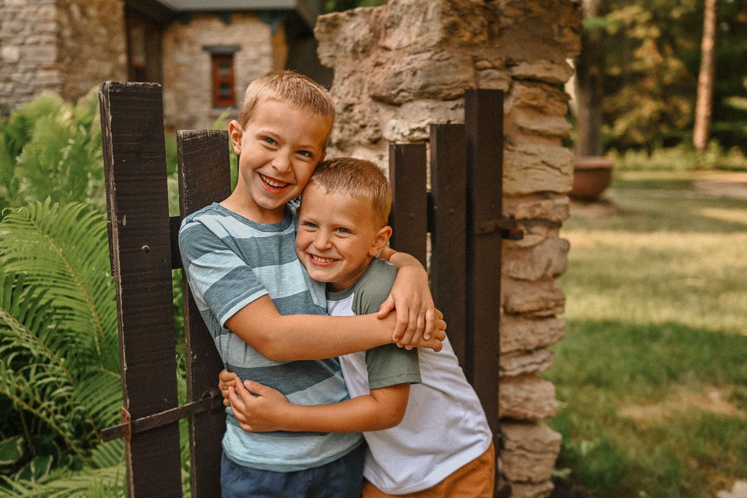 Two children embrace and smile while standing by stone pillar and wooden gate.
