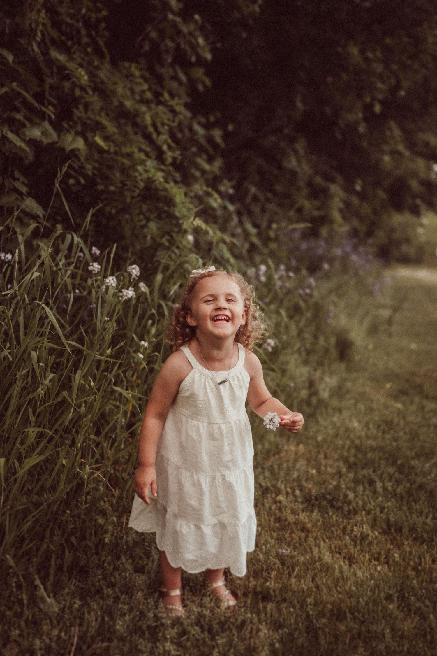 A candid portrait of a laughing child in a white dress standing among tall wildflowers at golden hour.