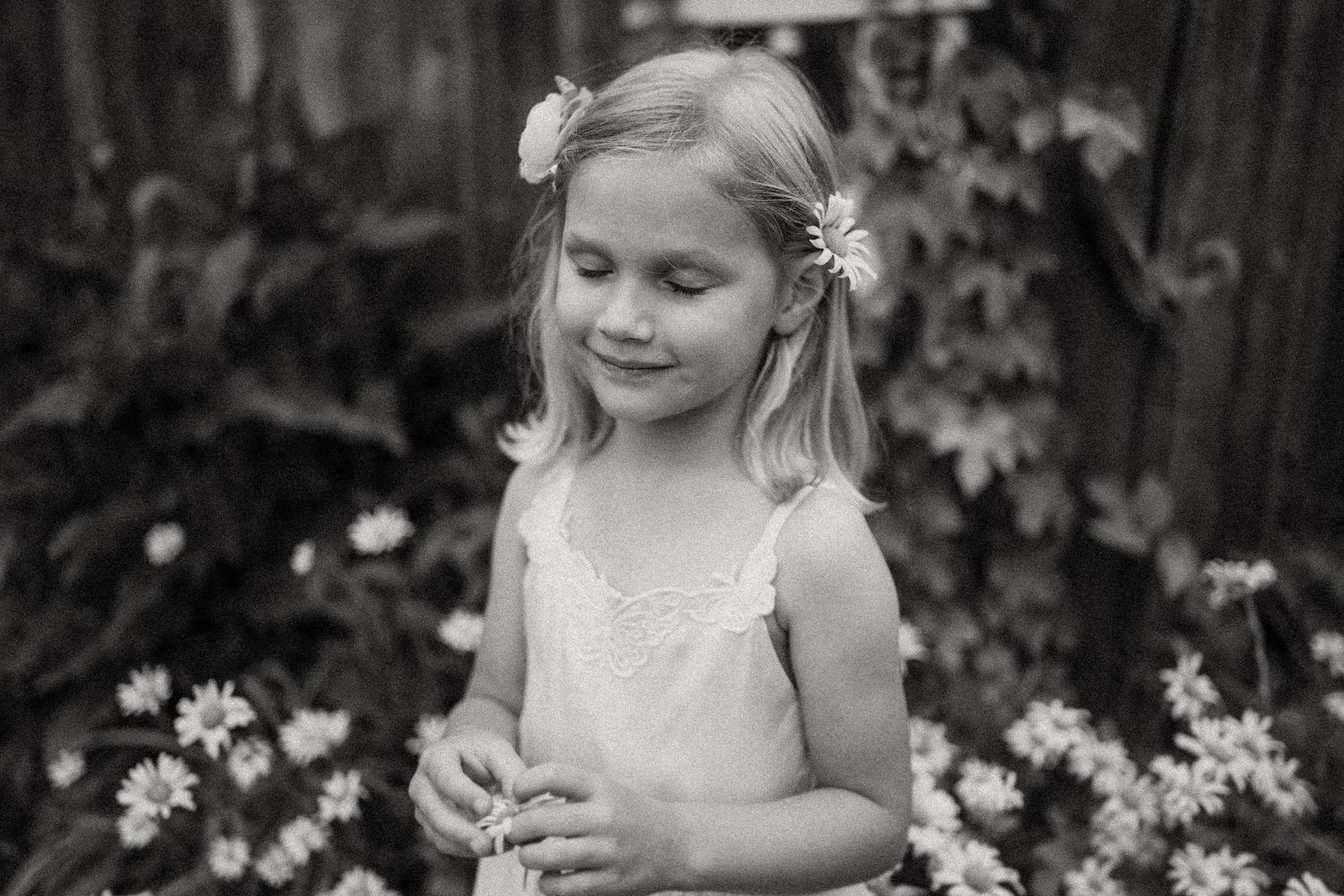 Black and white portrait of child in white dress standing among garden flowers.