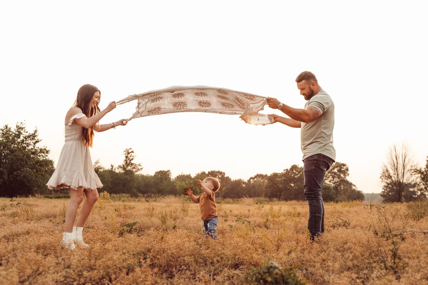 A family plays with a blanket in a golden field at sunset making an arch over a small child.