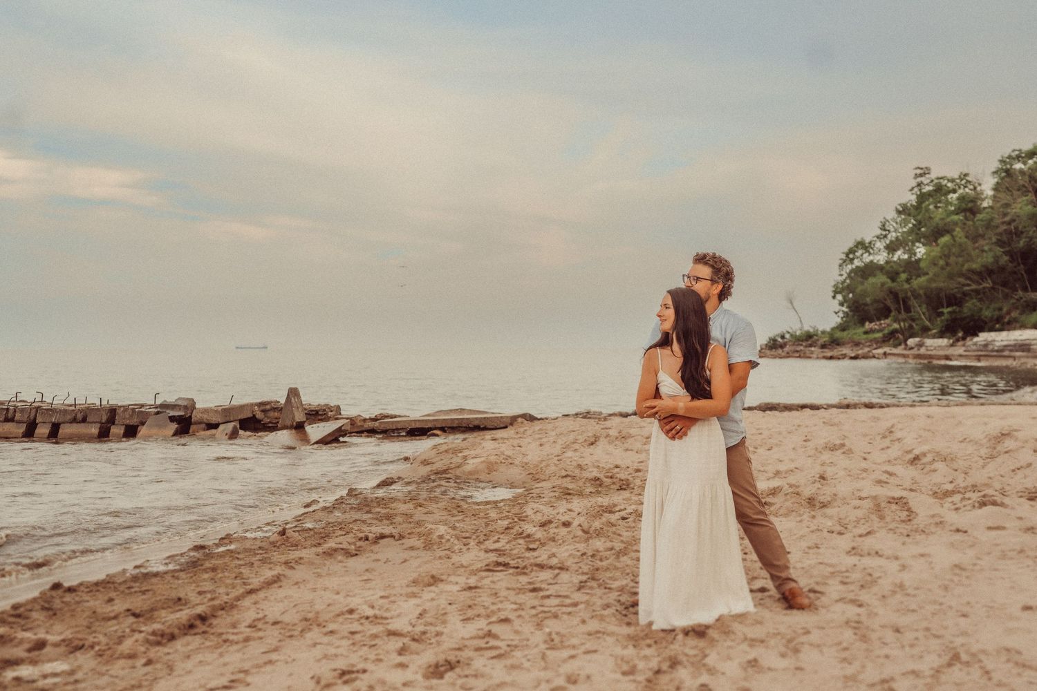 A couple shares a romantic moment at sunset on a beach with rock formations and trees in the distance.