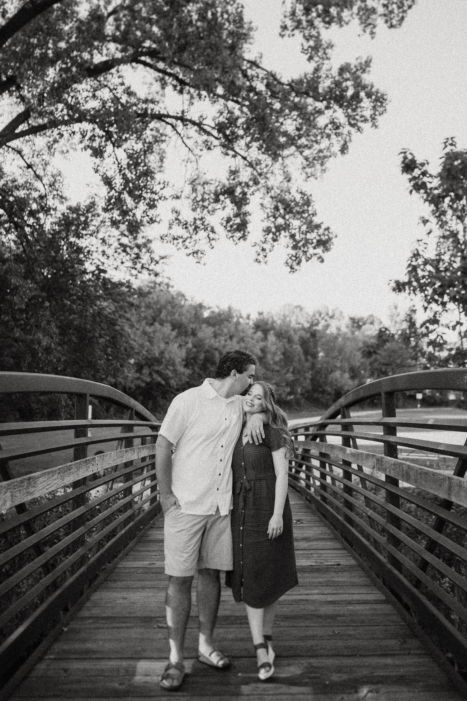 Black and white image of couple walking away on wooden bridge surrounded by trees.