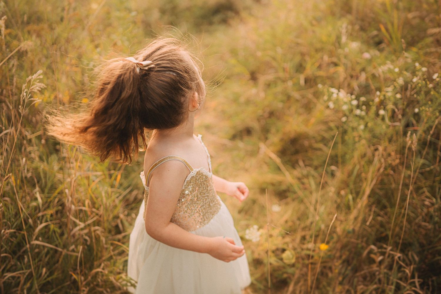 Child in sparkly dress dancing through tall grass in warm evening light.
