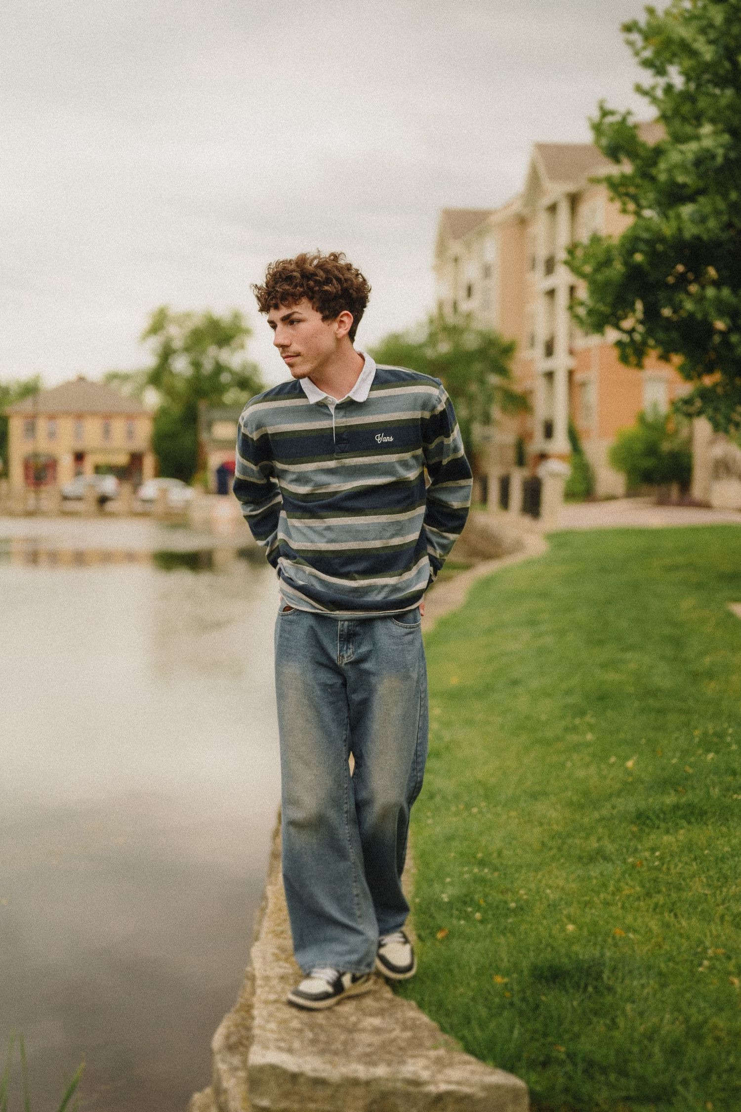 Someone wearing a striped sweater and jeans stands on a stone wall beside a lake near residential buildings.