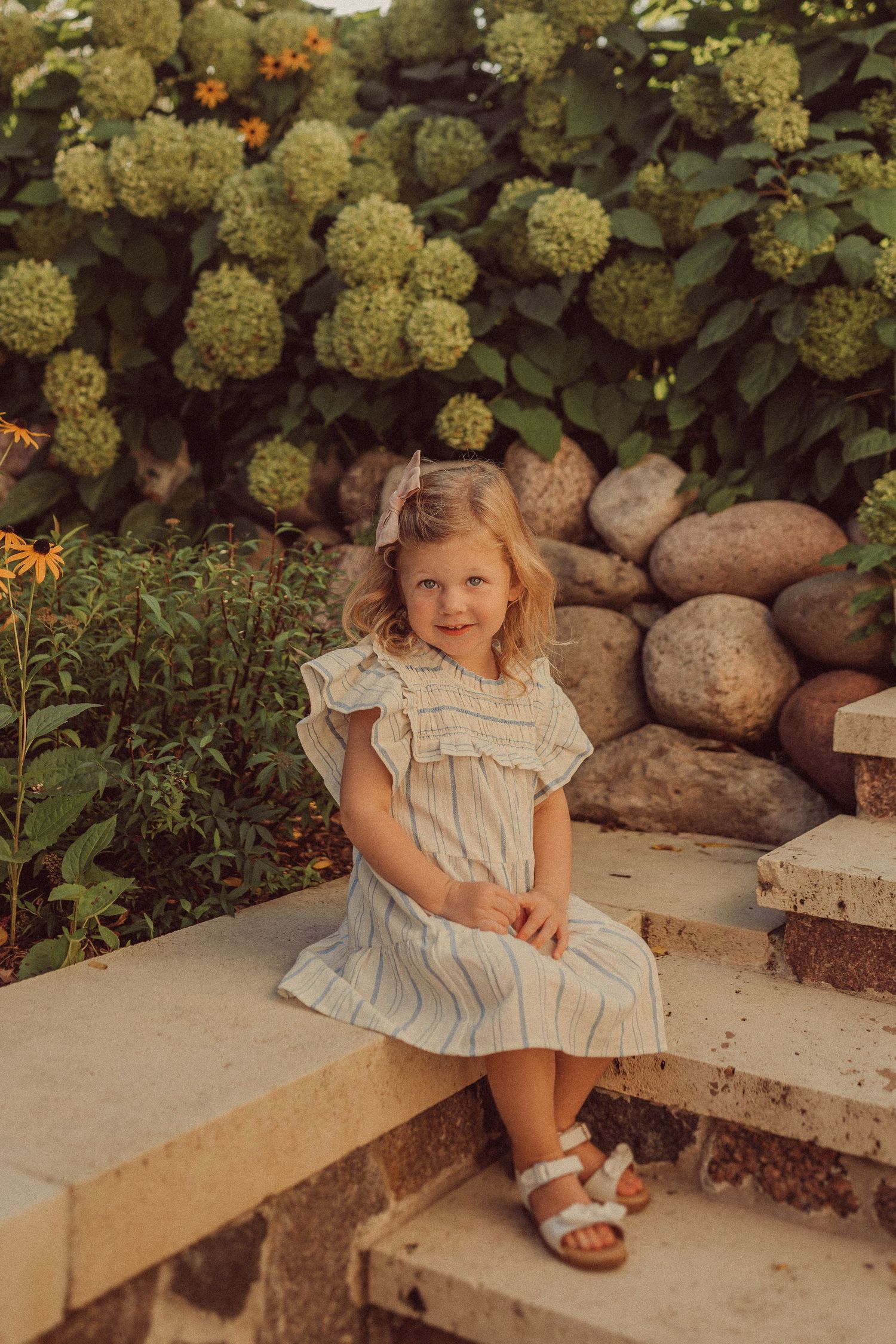 A child sits on stone steps wearing a white ruffled dress against a backdrop of hydrangea bushes and rocks.