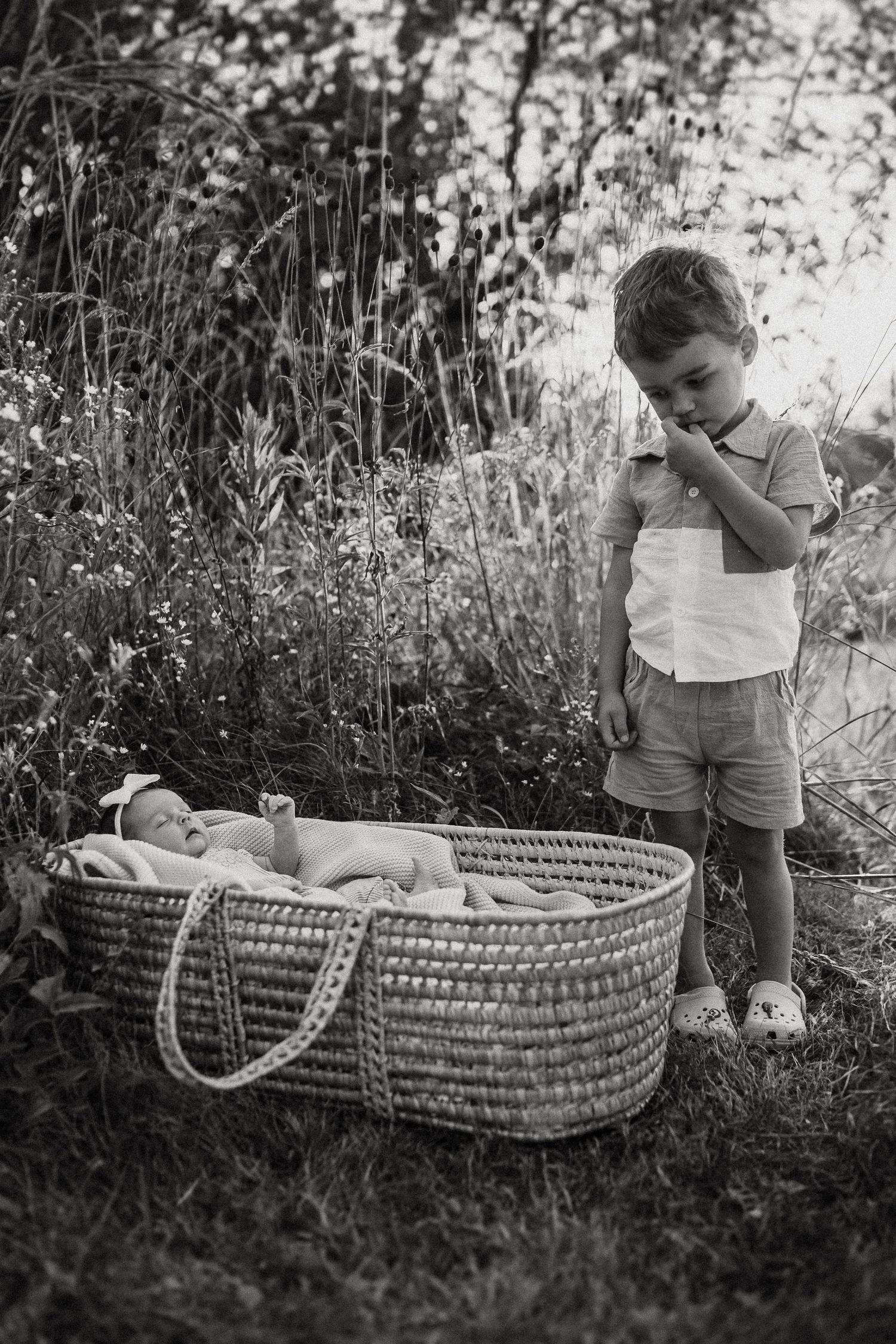 Young child standing next to wicker Moses basket in black and white outdoor portrait.