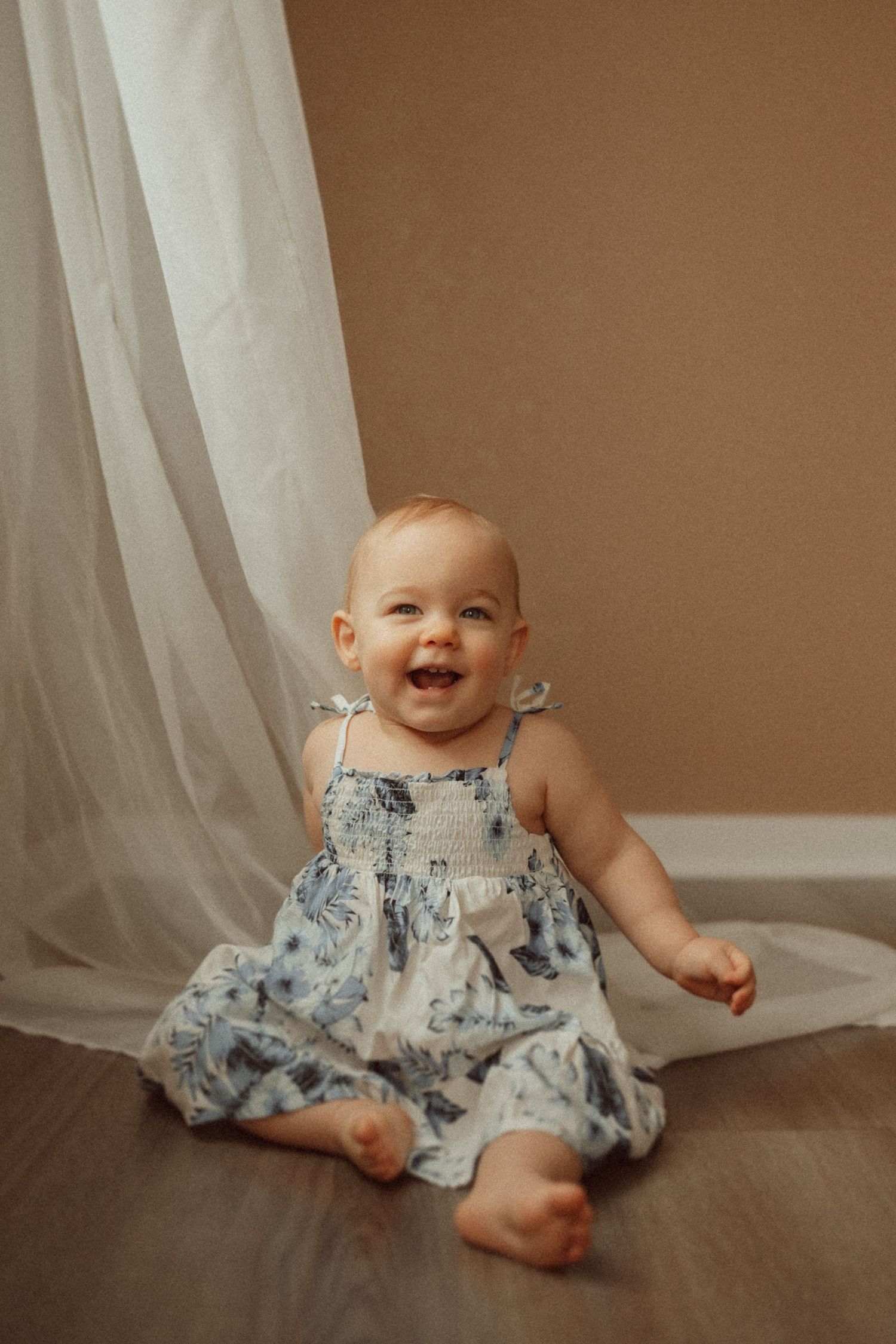A cheerful toddler in a floral sundress sits on wooden flooring near white curtains.