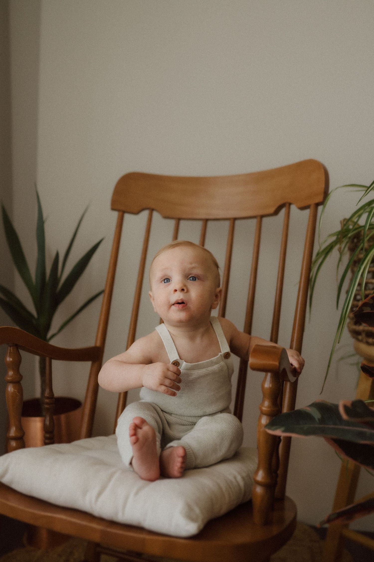 Baby in beige romper sitting on cushioned wooden rocking chair near indoor plants.