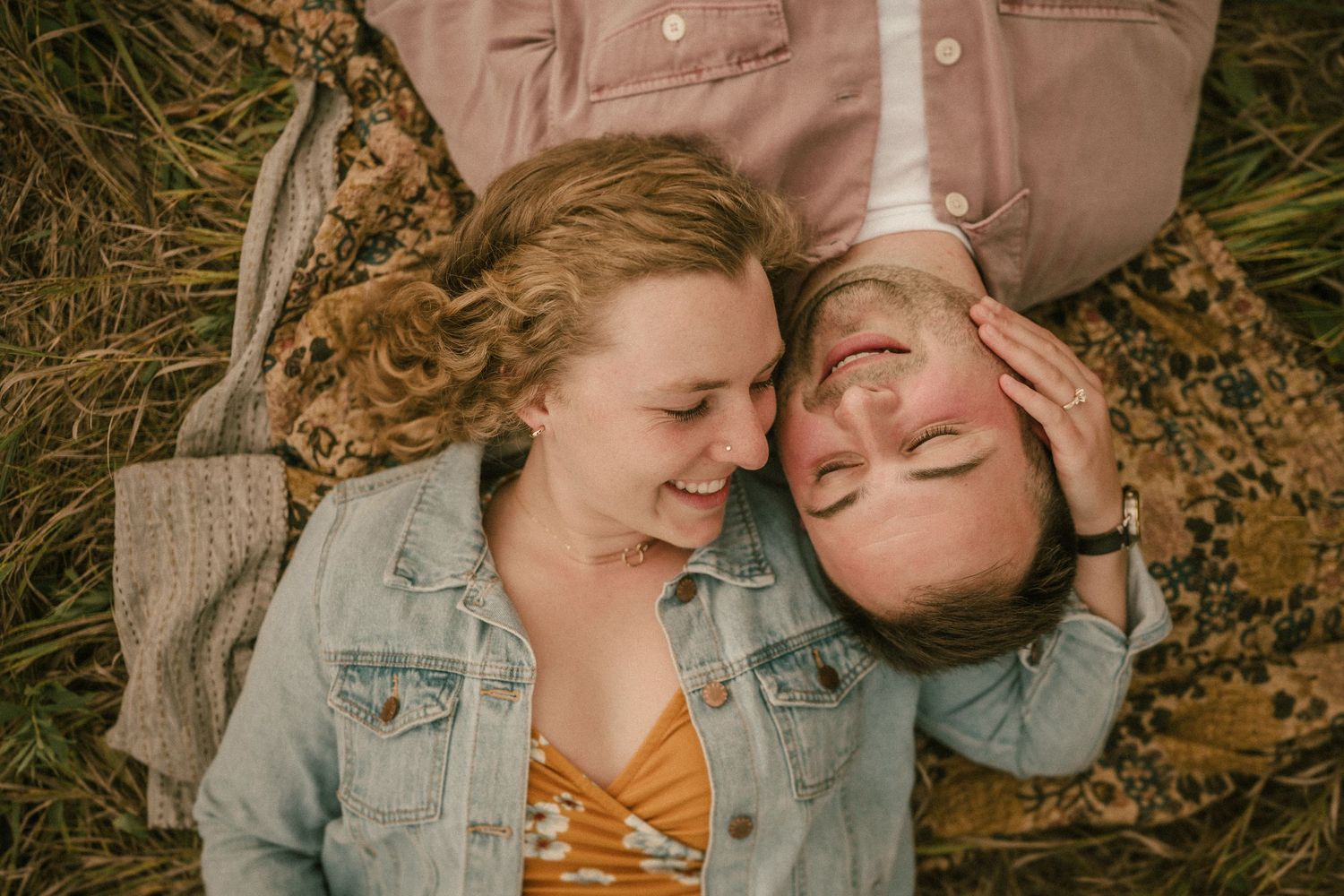 A couple lies head to head on a blanket outdoors sharing a tender moment.