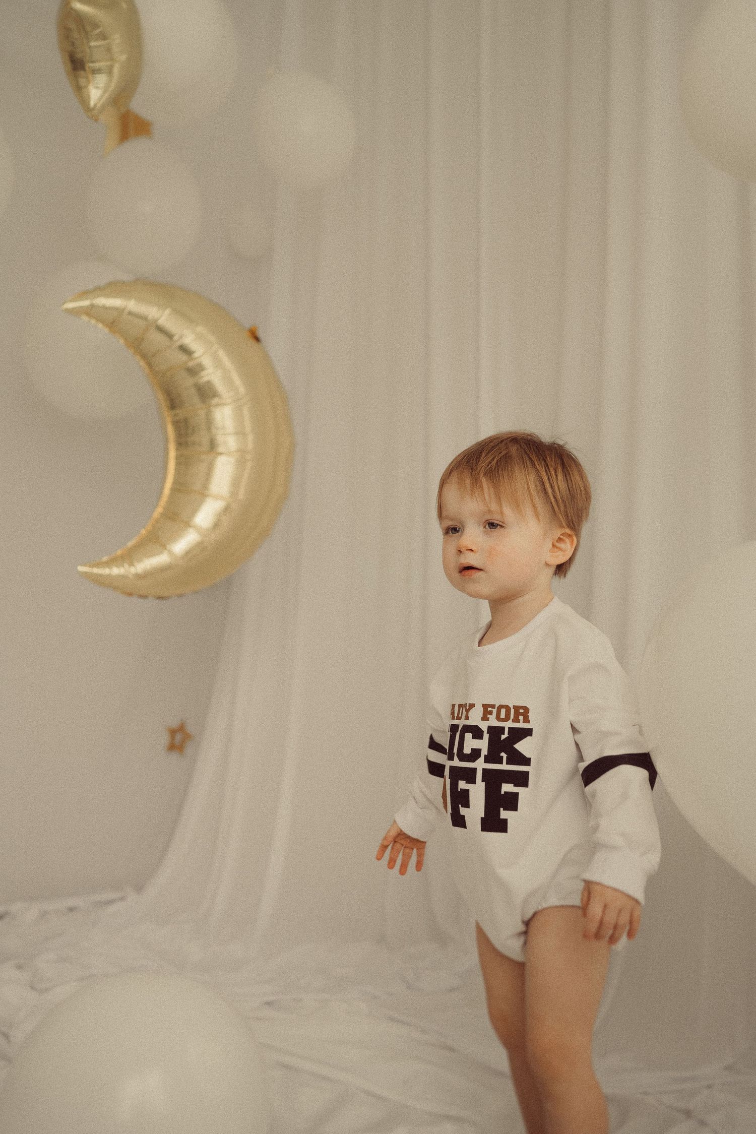 Toddler in white 'Kick Off' onesie standing against white backdrop with golden moon balloon decor.