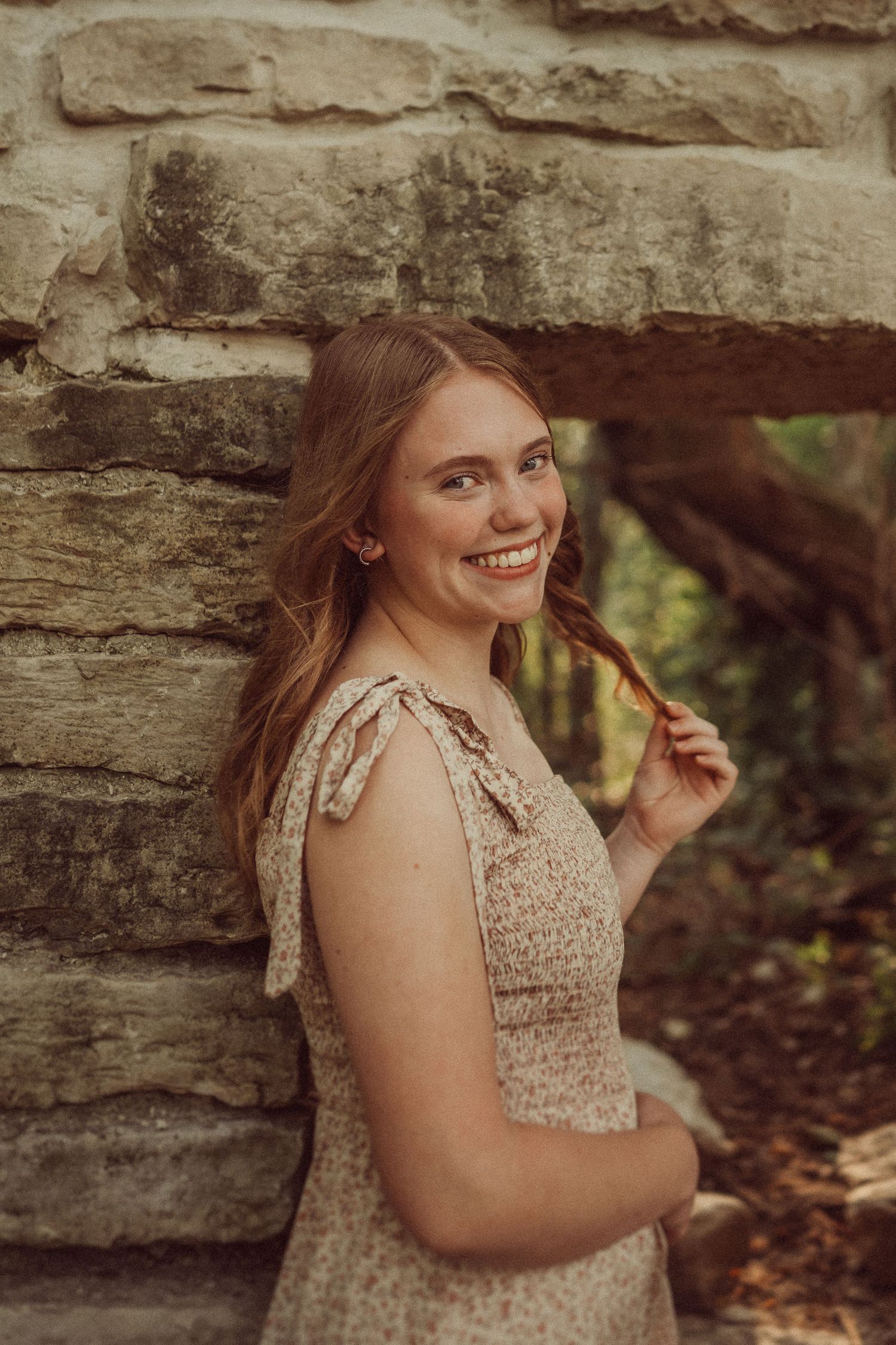 A smiling woman in a beige floral dress poses against a stone wall in a natural outdoor setting.
