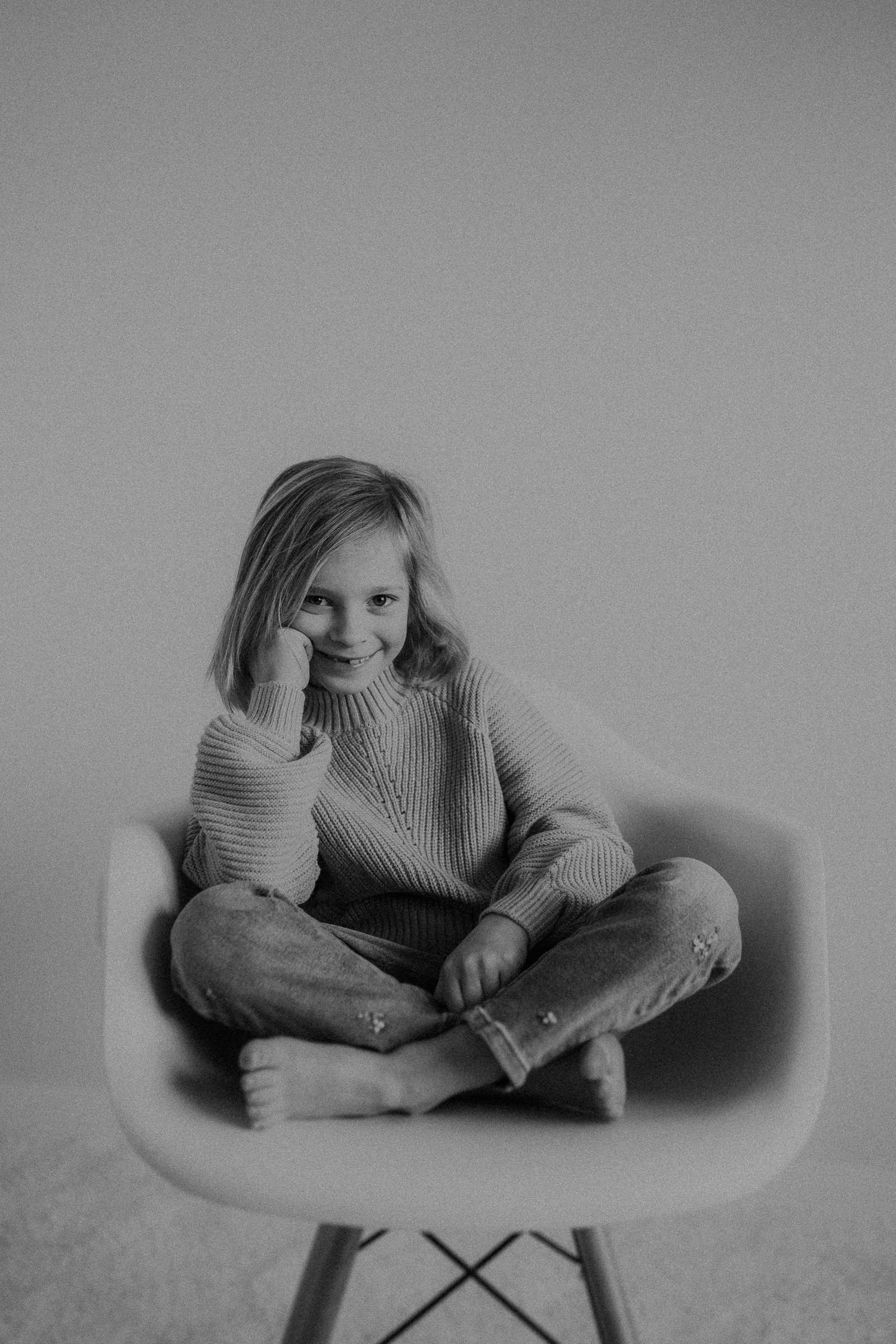 Child in ribbed sweater sits cross-legged on white modern chair in black and white photo.