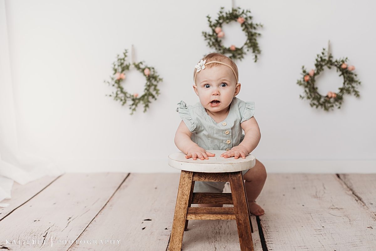 Baby in white top crawling on wooden floor with floral wreaths on wall.