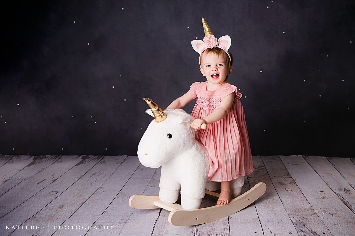 An excited toddler in pink plays on a white rocking unicorn toy against a dark backdrop.