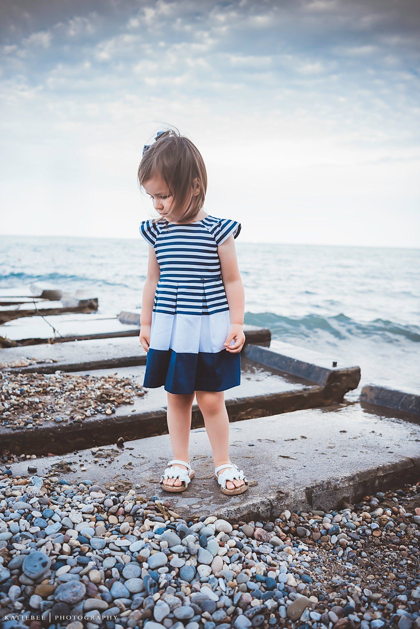 A small child in nautical dress stands thoughtfully on a rocky beach beside the ocean.