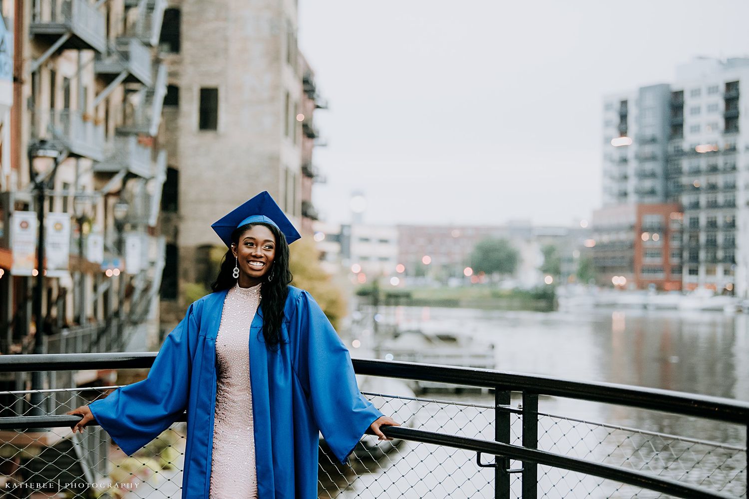 Graduate in blue cap and gown standing on waterfront with city buildings in background.
