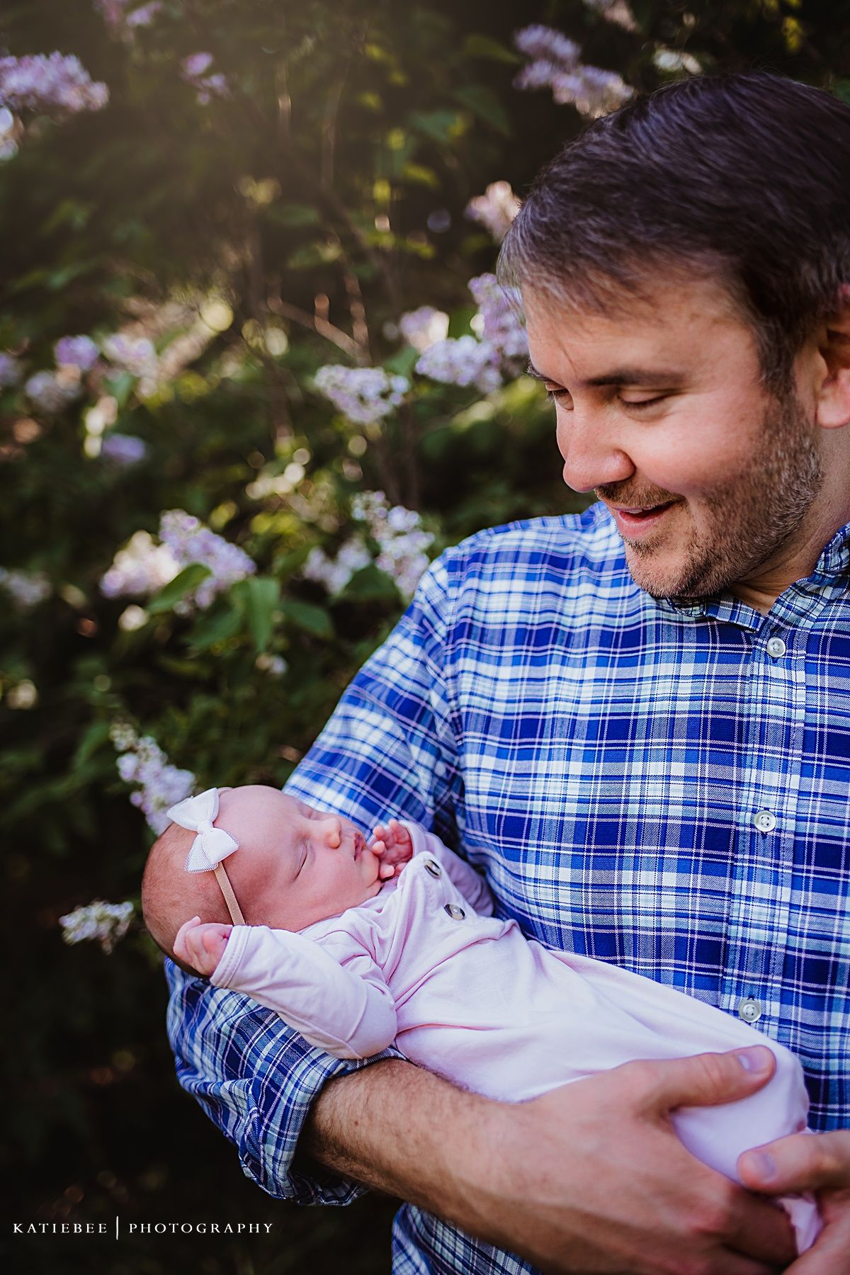 Tender outdoor portrait with flowering bushes as a tiny baby rests against a blue plaid shirt.