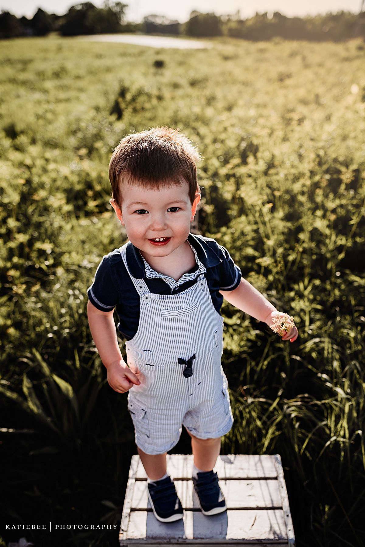 Toddler in white overalls and navy shirt smiling while standing on wooden platform in grassy field.