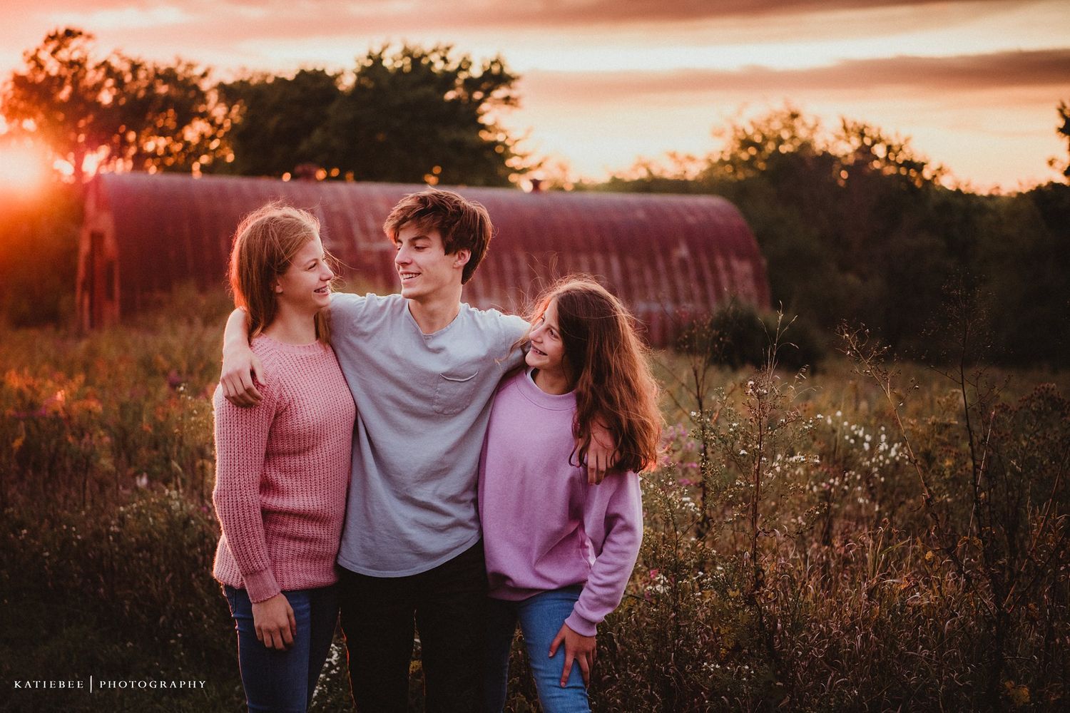 Three people embracing and laughing together at sunset with a red barn in background.