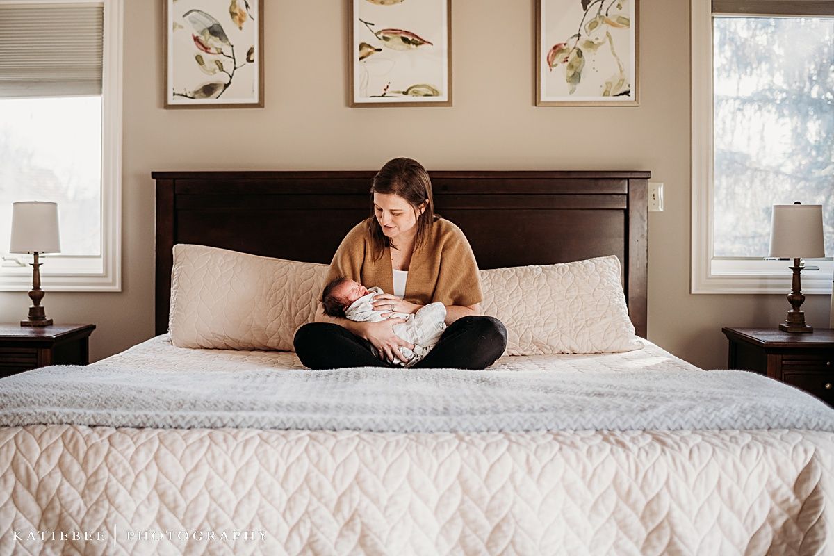 Person in tan sweater sitting cross-legged on bed with white quilt beneath nature artwork.