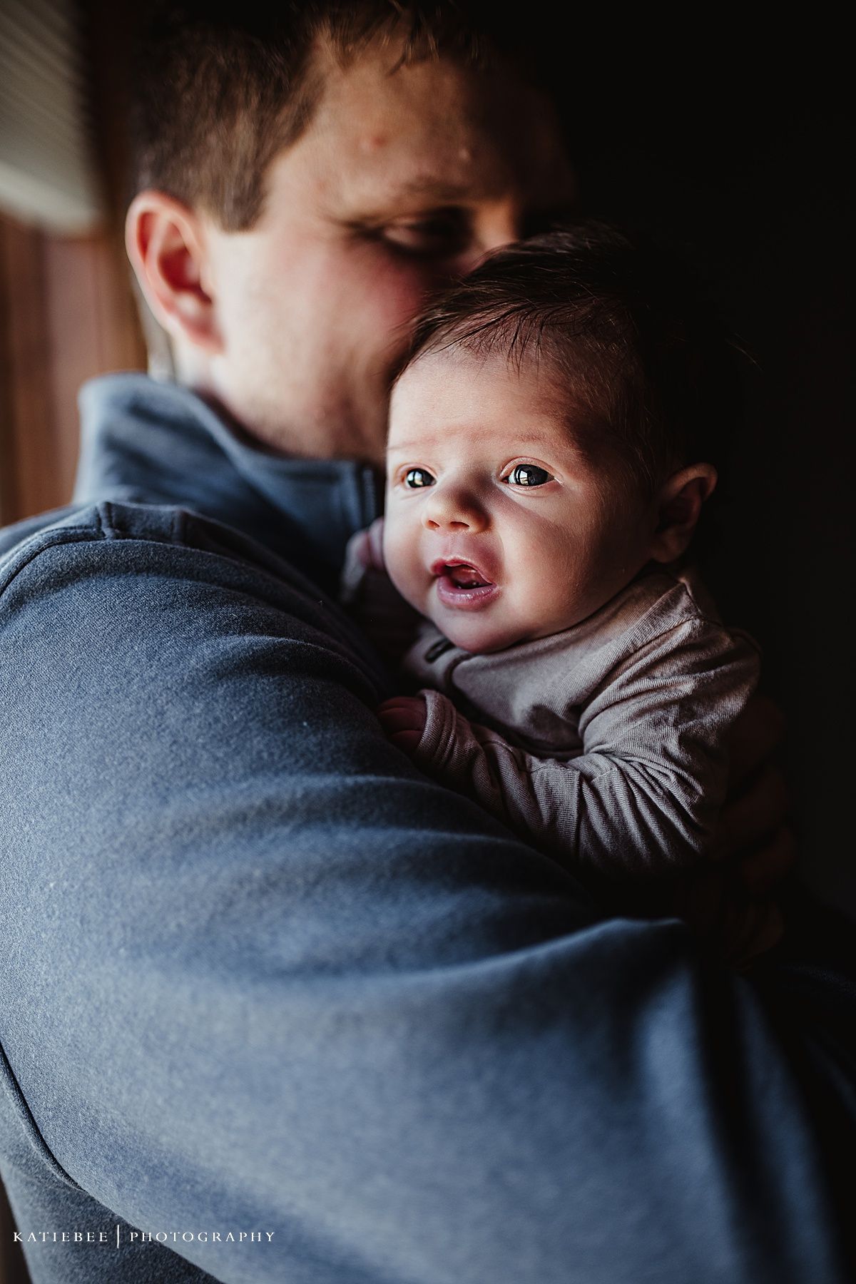 Sweet candid moment of baby in grey outfit being held against grey sweater in dim lighting.