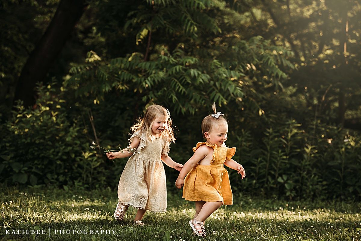 Two children in flowy dresses playing and running together in a sunlit garden.