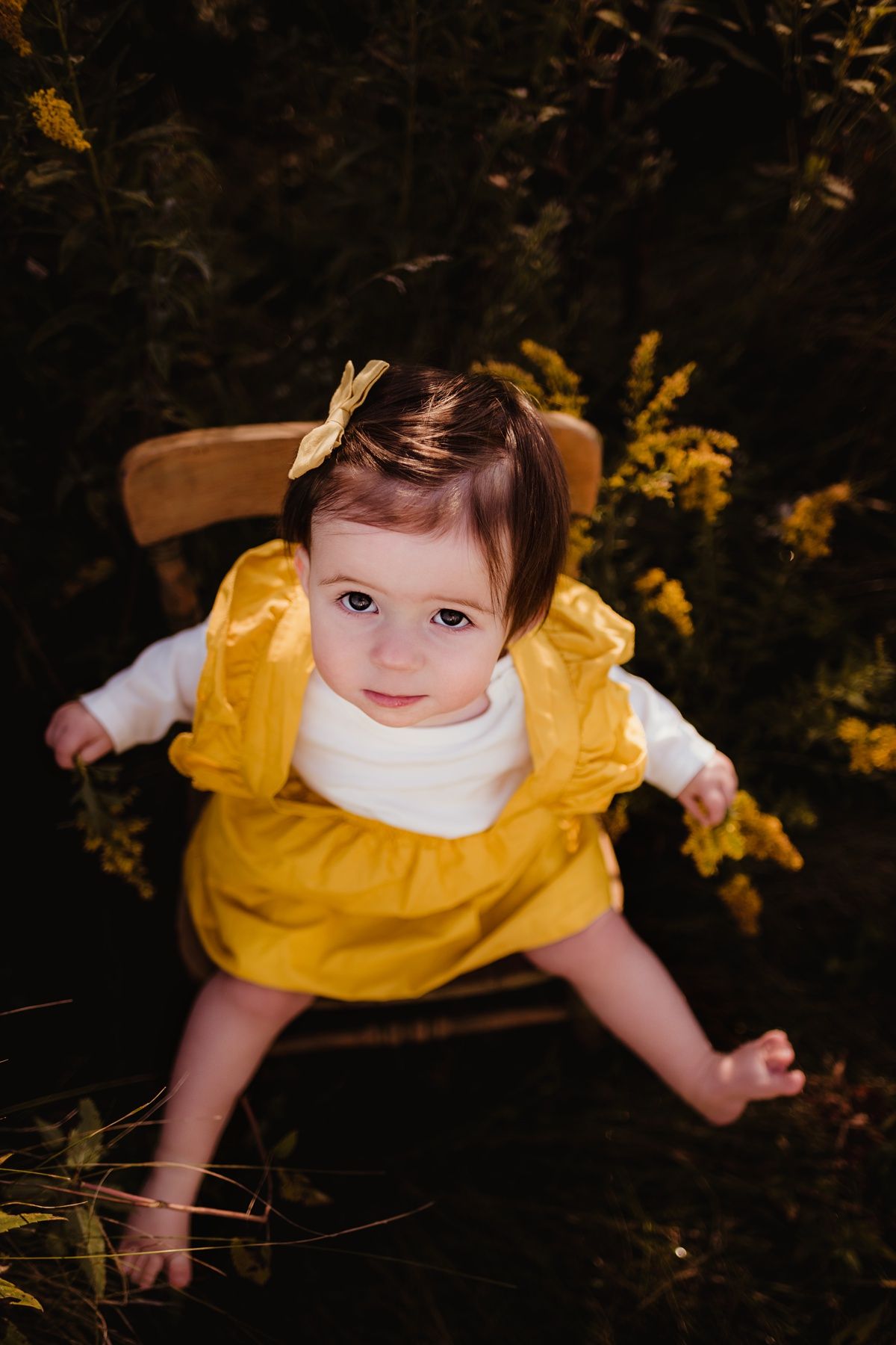 A small child in a yellow dress sits on a wooden chair surrounded by autumn foliage.