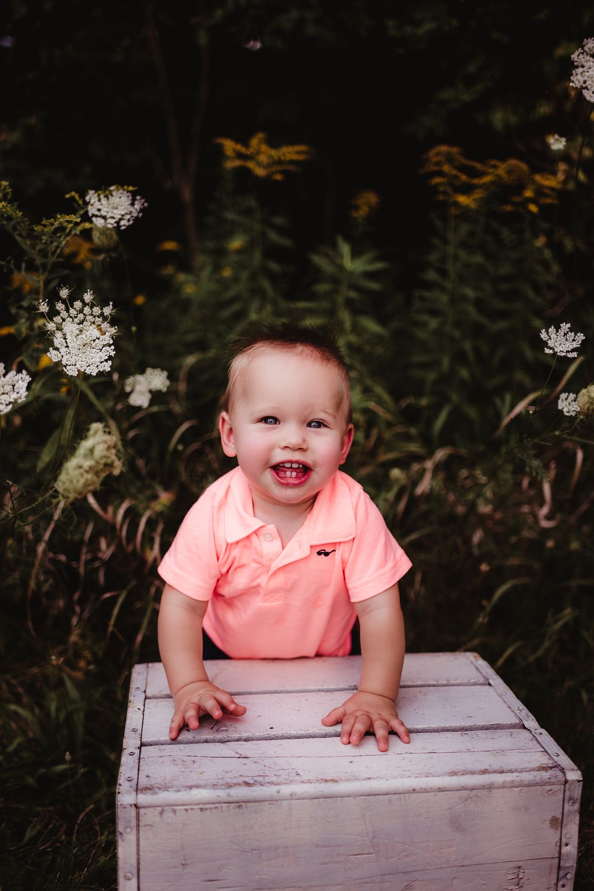 Baby in coral shirt smiling while leaning on white wooden crate in garden setting.