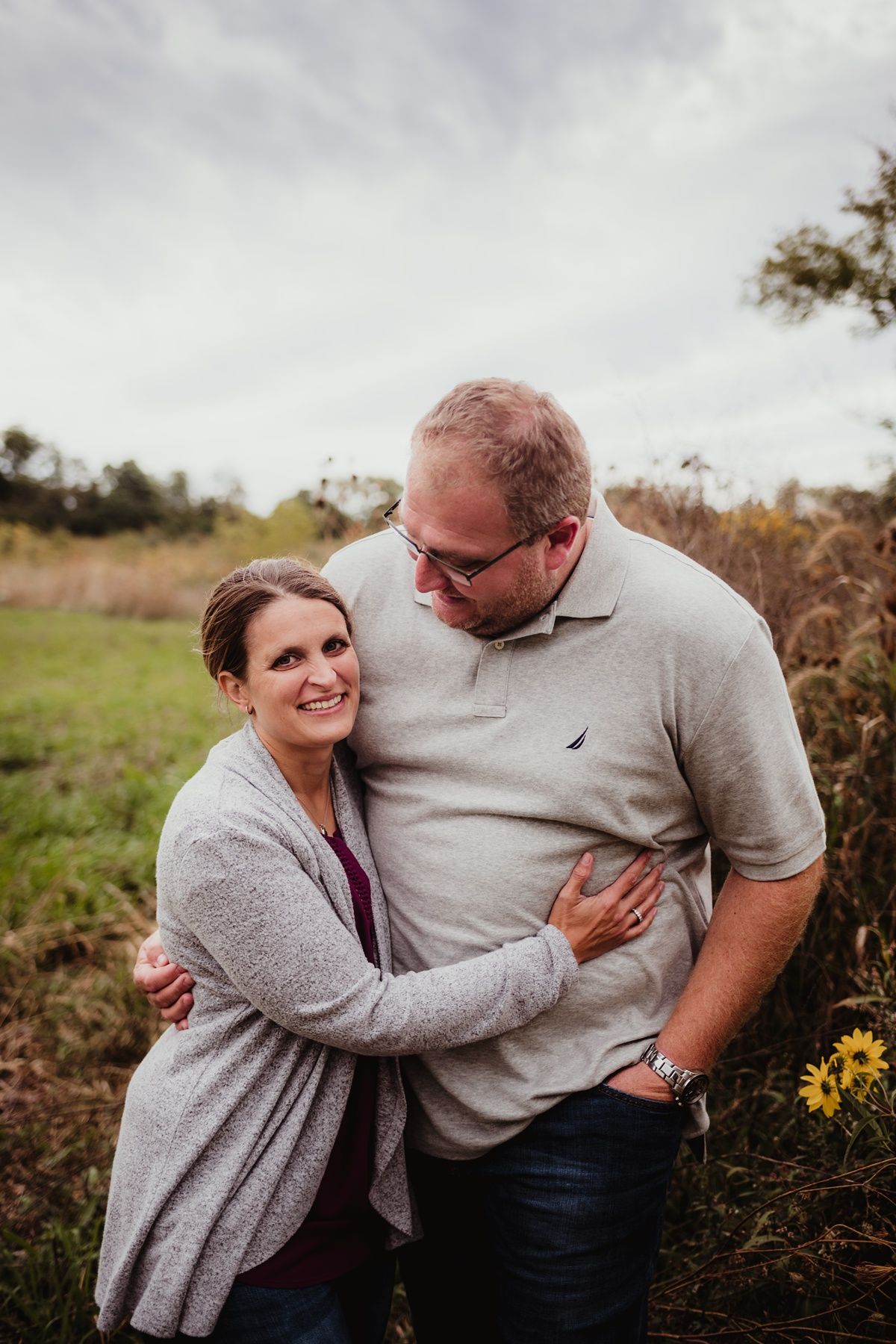 A couple shares an intimate moment outdoors while wearing matching gray sweaters in a field with wildflowers.