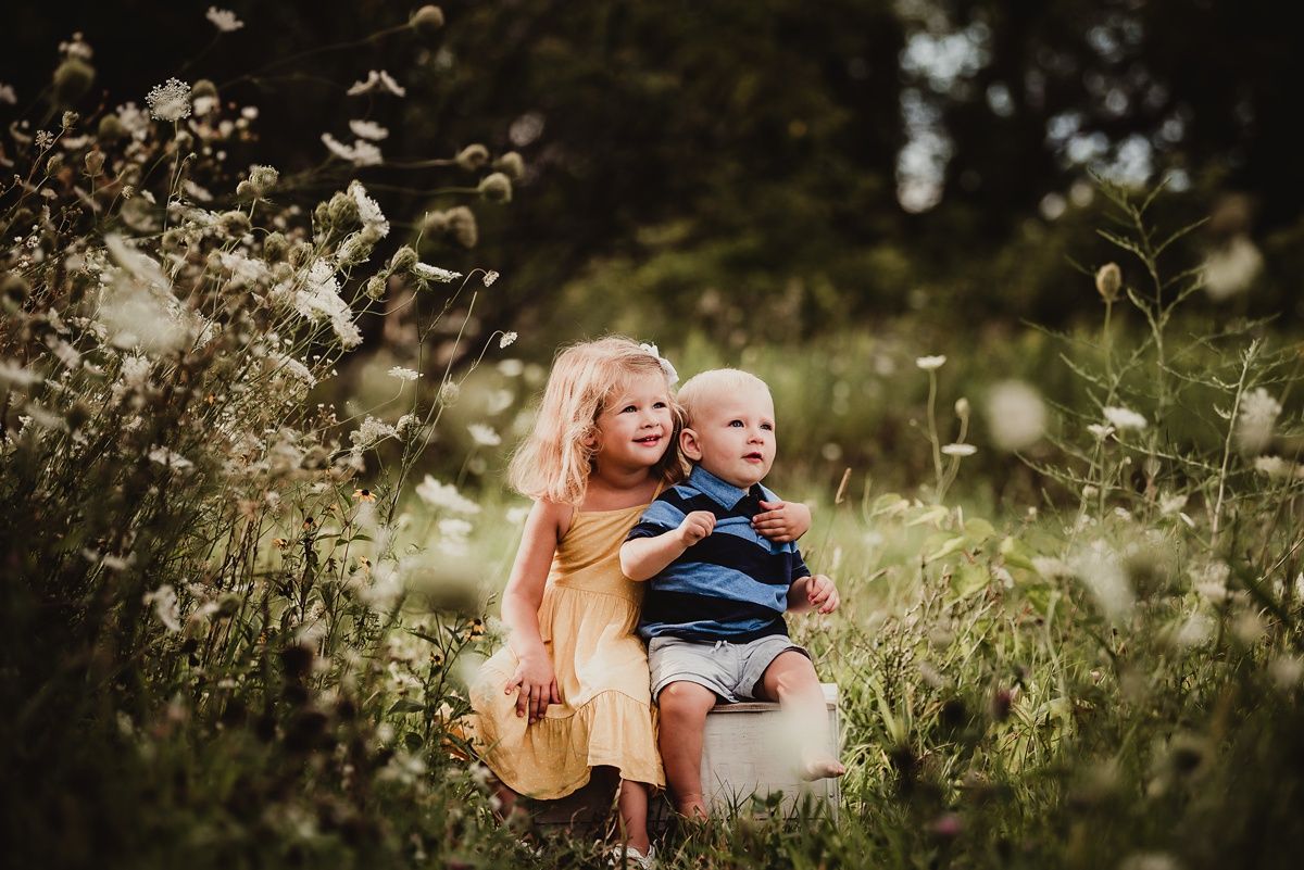 Two children sitting on a rustic stool in a meadow filled with tall grass and wildflowers.