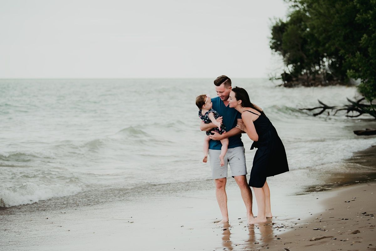 A family shares a tender moment while standing at the edge of the ocean on a cloudy beach day.