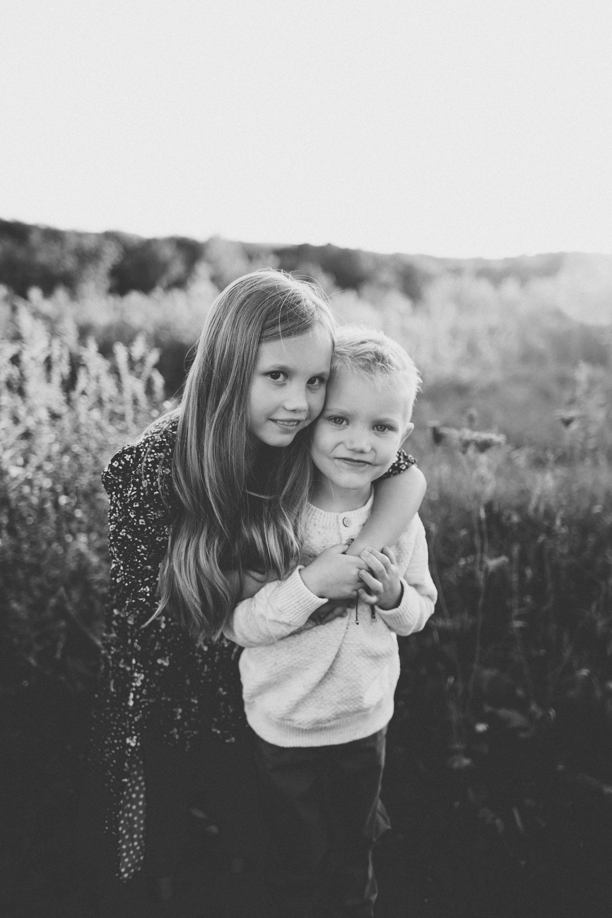 Black and white portrait of siblings hugging in field of wildflowers.