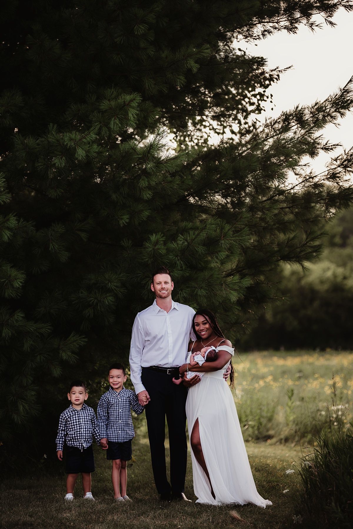 A family stands together under pine trees in a meadow at sunset for a portrait.