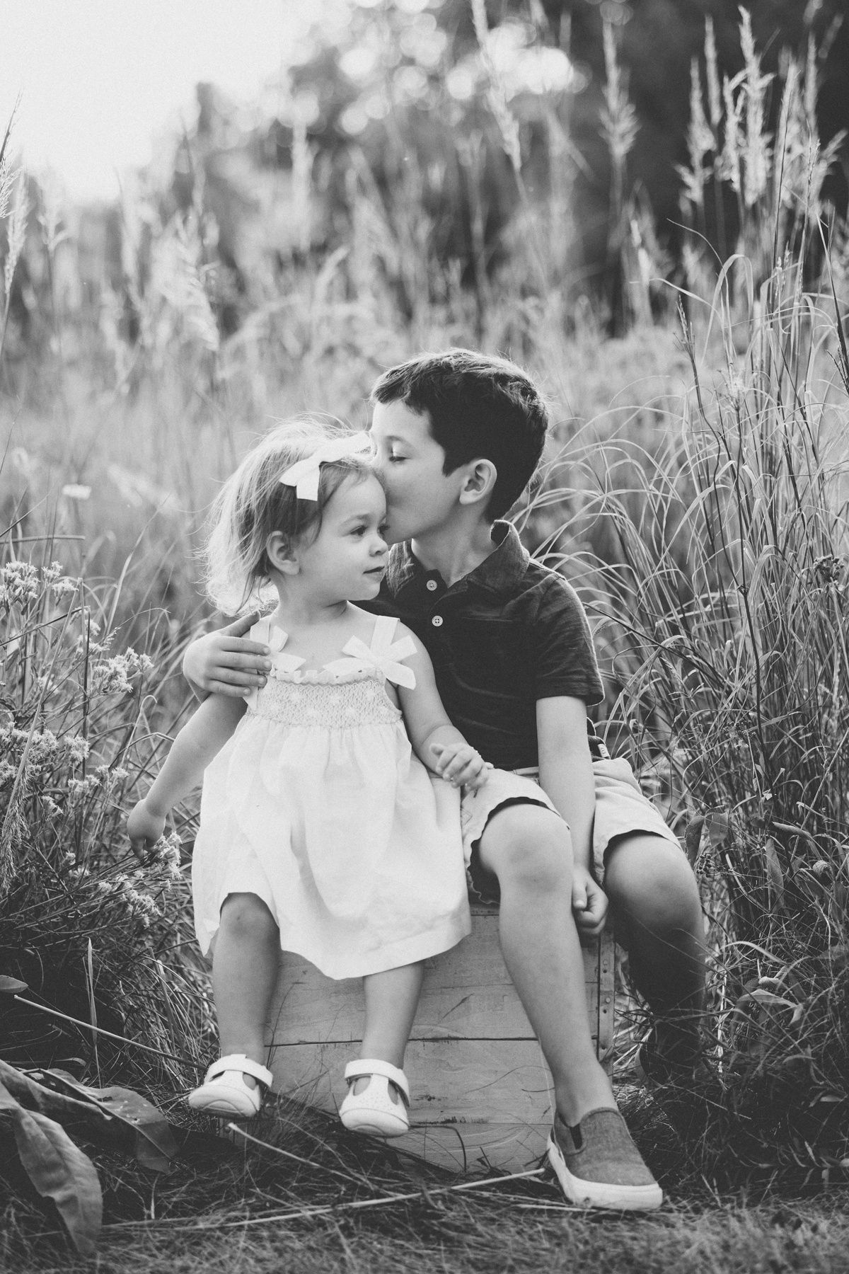 Black and white photo of two children sharing a sweet moment while sitting among tall grass.