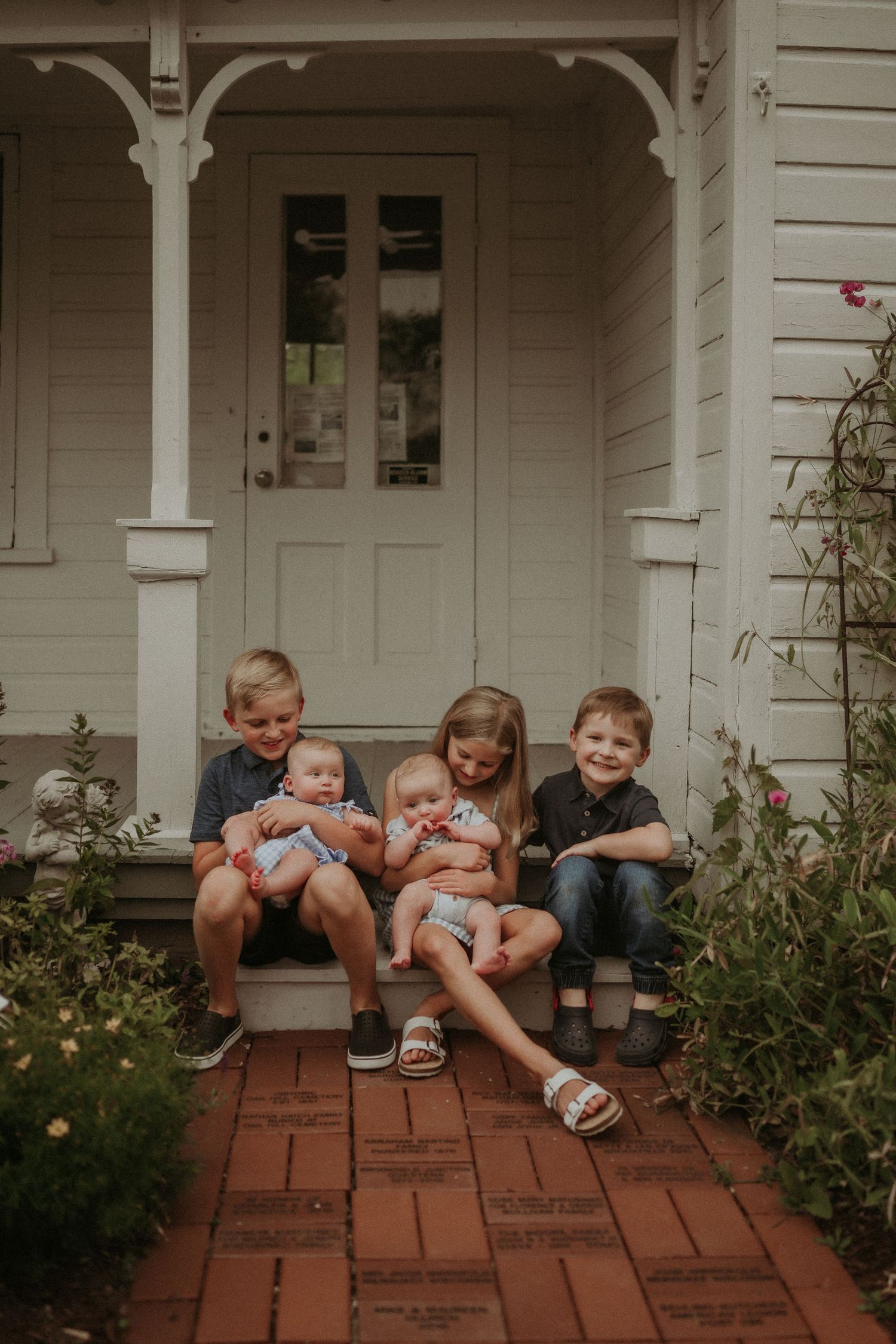 Children sitting on brick steps of farmhouse porch with two infants in casual outdoor setting.