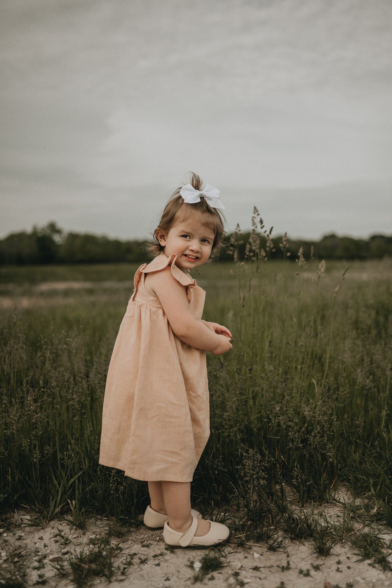 A small child in a peachy dress poses playfully in a wild grassy field.
