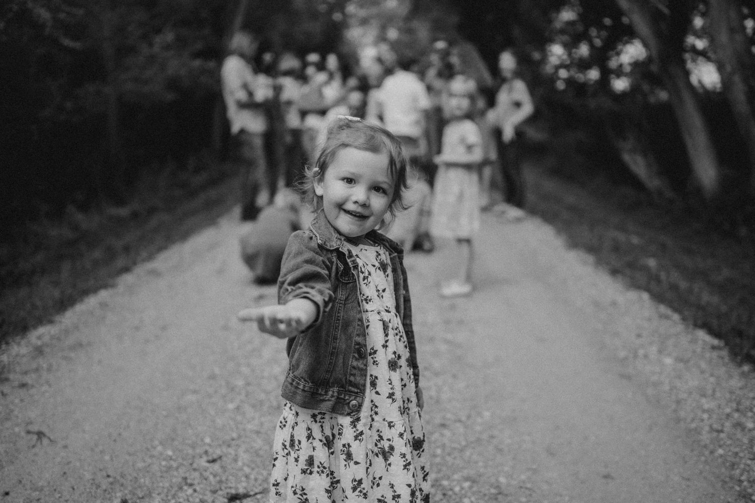 Child in a dress and jacket poses on a gravel path with others visible in background.