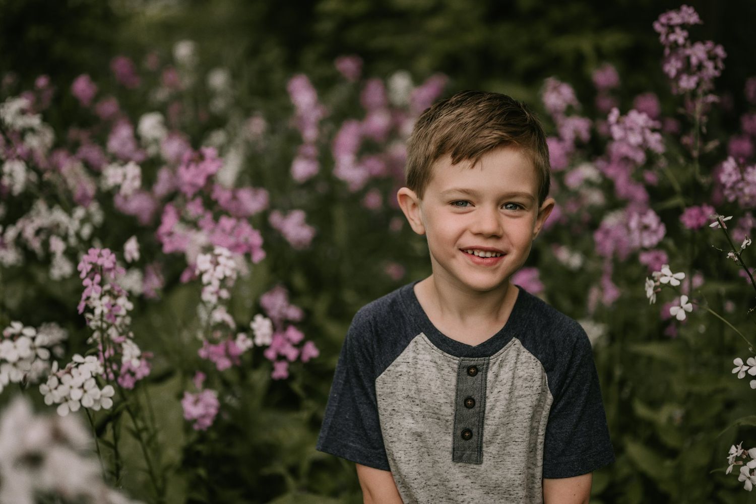 Child poses in a gray henley shirt against blooming pink wildflowers in a garden setting.