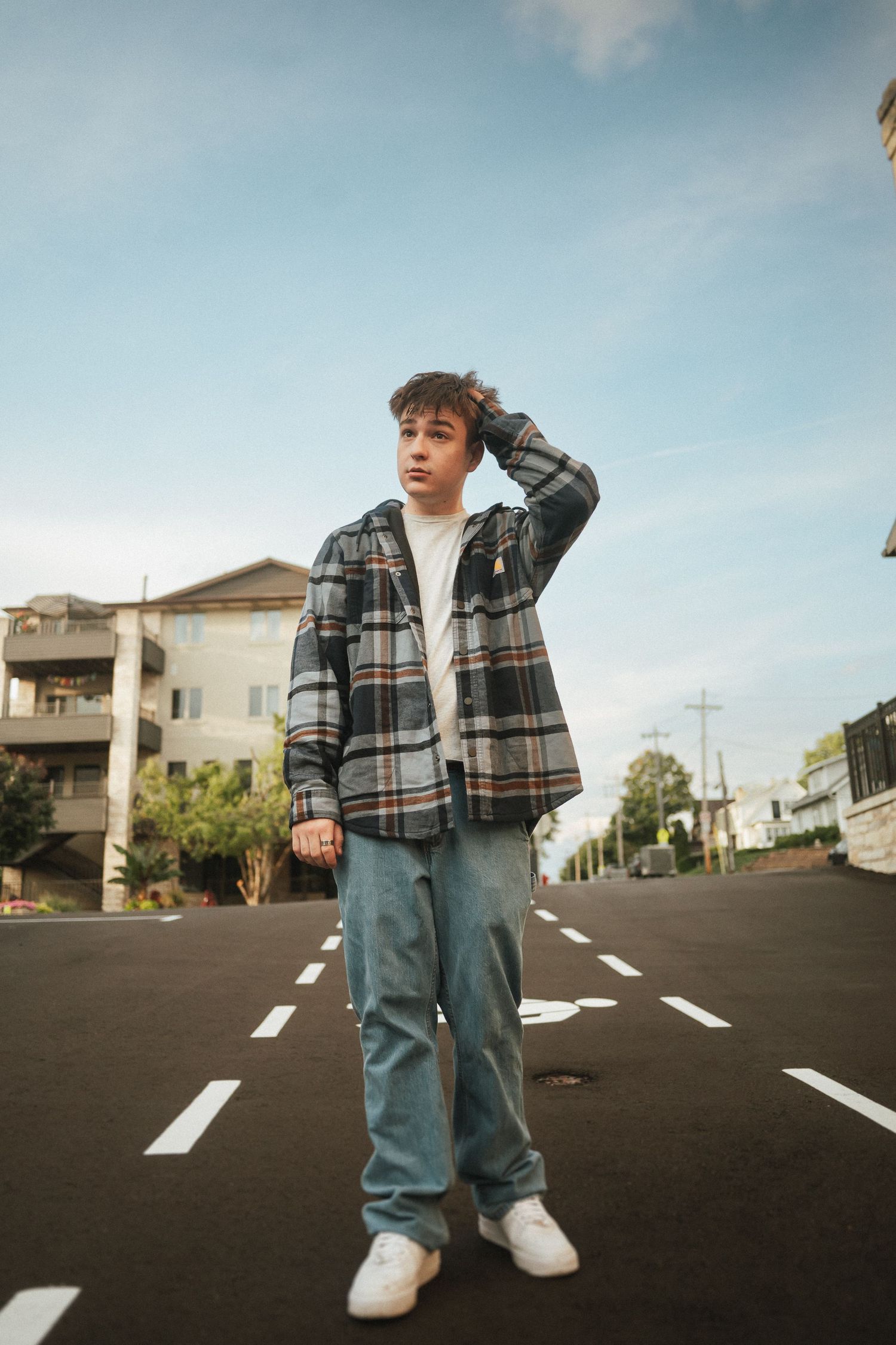Person in plaid jacket and jeans standing on empty street with white road markings and buildings in background.