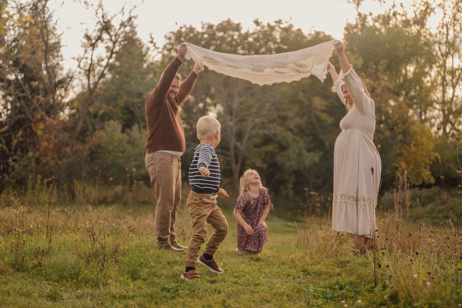 Family enjoying a playful moment in a sunlit meadow with autumn trees in background.