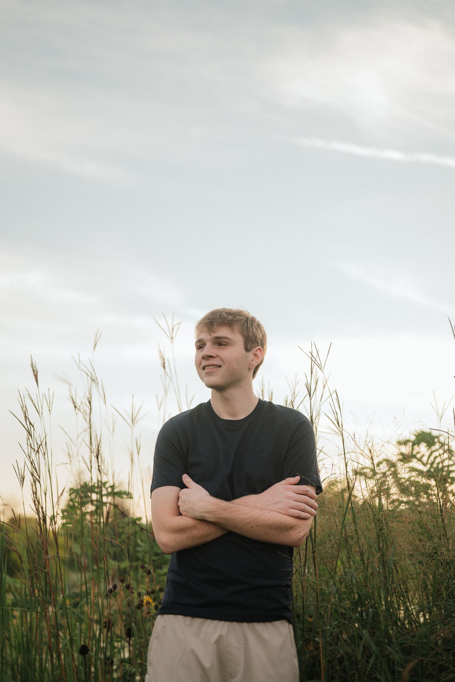 Person in black shirt standing in tall grass field against cloudy sky during golden hour.