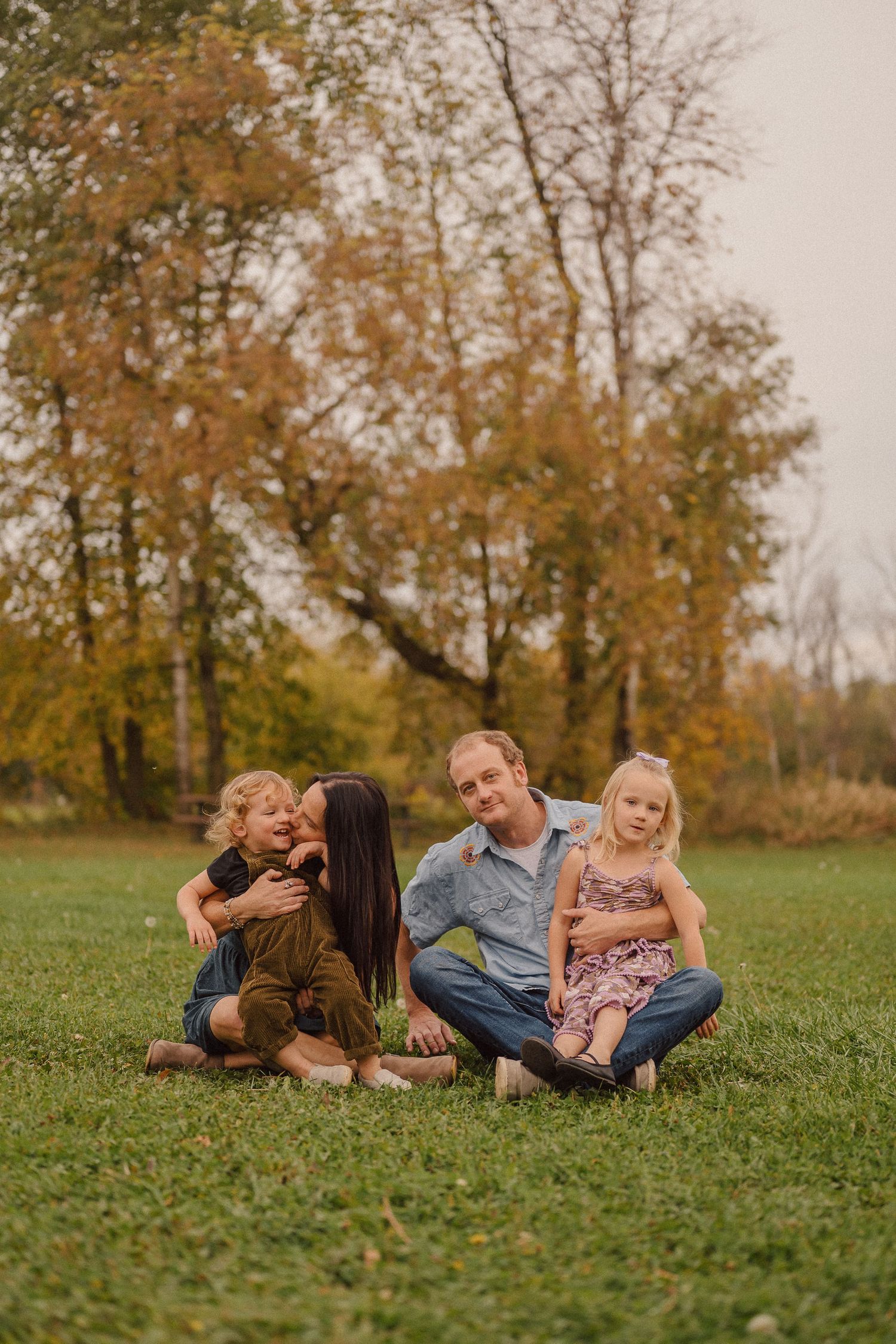 A relaxed outdoor family scene on green grass with autumn trees creating a warm golden backdrop.