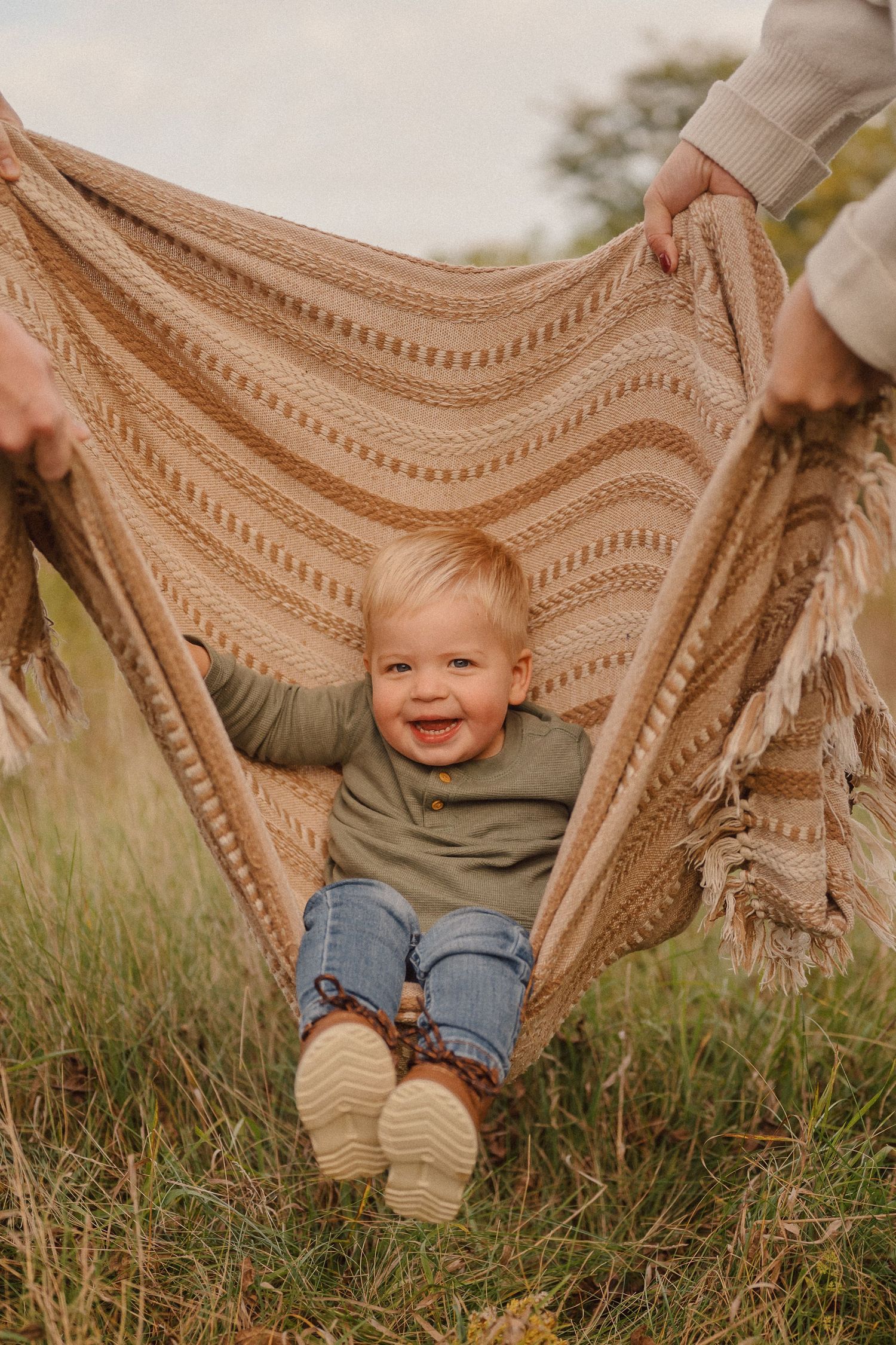 A beige knit blanket is held up in a grassy field during sunset creating a cozy outdoor scene.