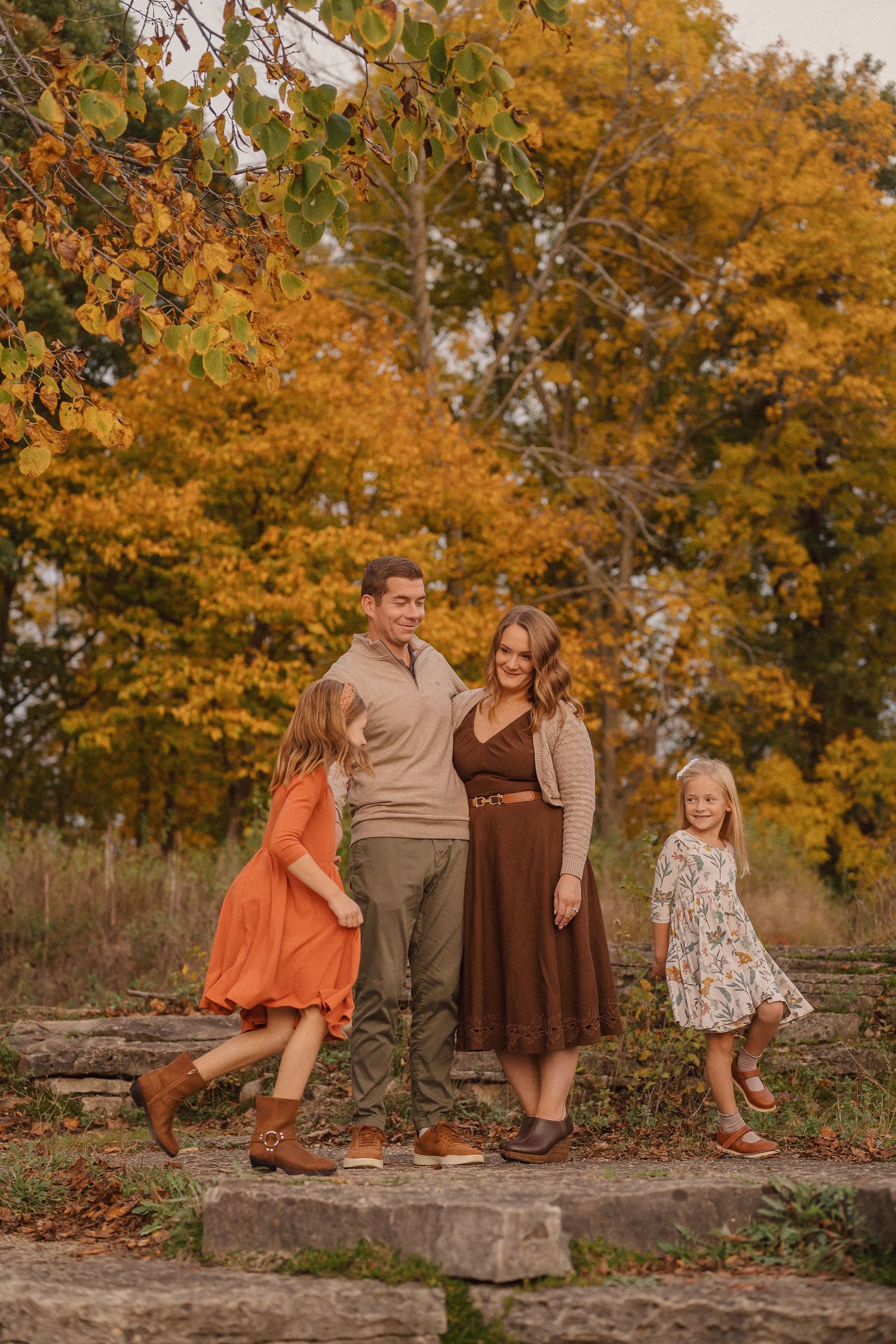 Family members pose casually on rustic stone steps with golden fall trees creating a warm backdrop.