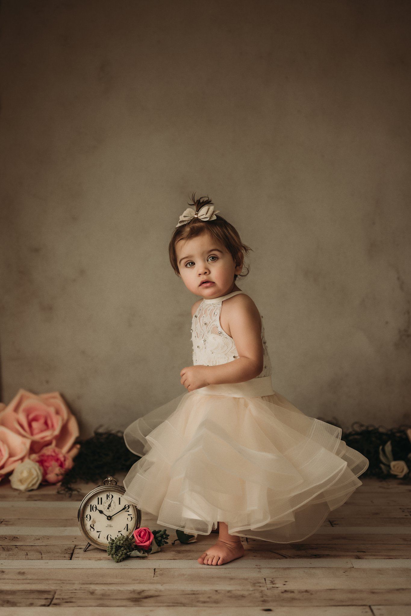 A delicate tulle dress adorns a tiny subject posing with vintage clock and roses on wooden flooring.