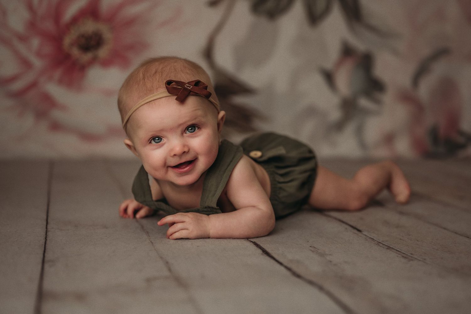 Child lying on wooden floor wearing olive romper and bow against floral backdrop.