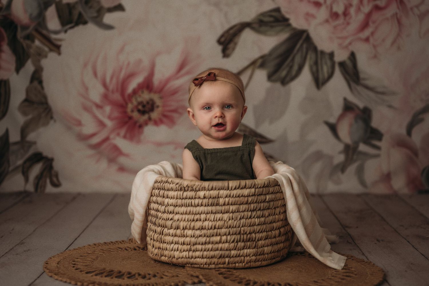 Baby in olive romper sitting in woven basket with white fabric.